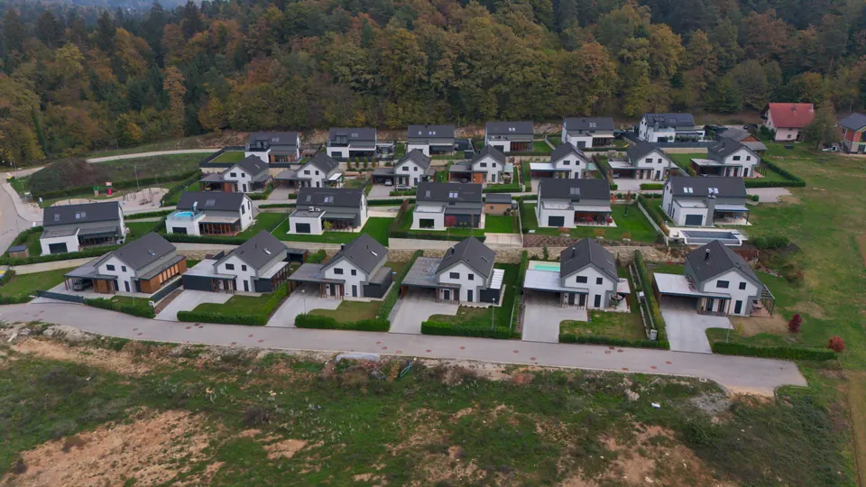 Vrsta novih belih hiš s temnimi strehami, nadstreški in urejenimi vrtovi ob ulici, posneta z dronom od spredaj, za njimi jesenski gozd