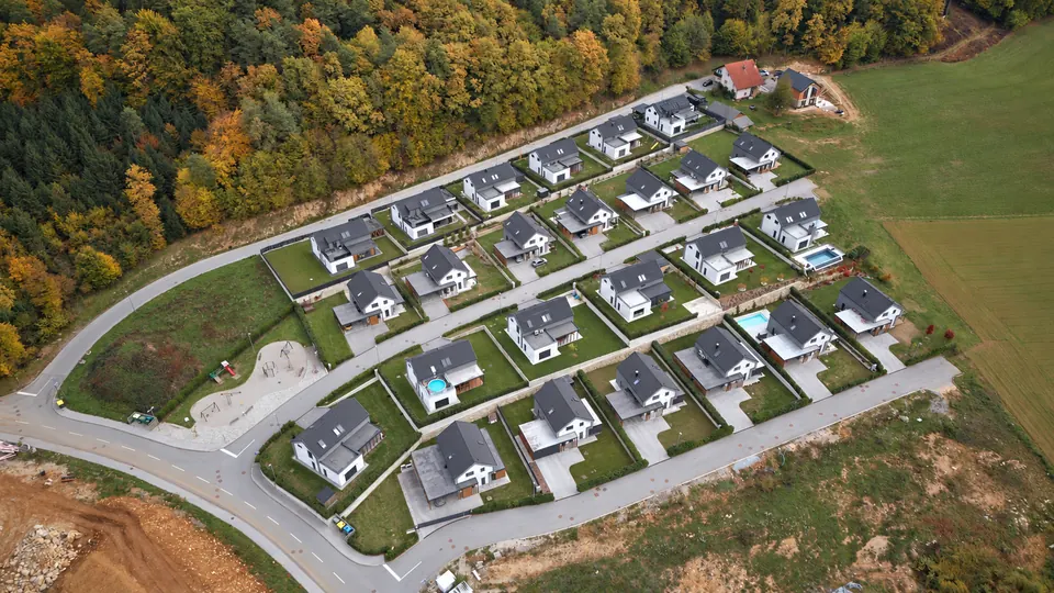 Novo naselje enodružinskih hiš s temnimi strehami, vrtovi, bazeni in otroškim igriščem, posneto z dronom od zgoraj ob jesenskem gozdu